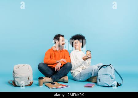 felici studenti multietnici seduti vicino a zaini e copybook e sorridenti l'uno sull'altro su sfondo blu Foto Stock
