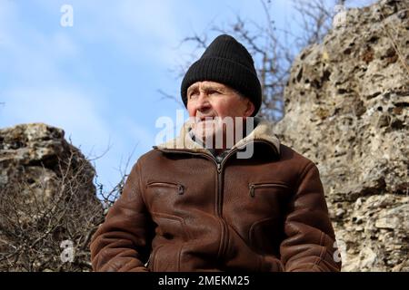 Ritratto dell'uomo anziano in pelle e cappello a maglia in montagna su sfondo cielo blu. Concetto di duro Highlander, di viaggio e di libertà Foto Stock