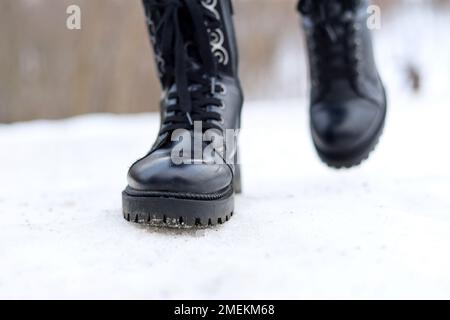 Gambe femminili in pelle nera stivali con lacci su un sentiero innevato. Donna che cammina su strada invernale, scivolosa in clima freddo Foto Stock