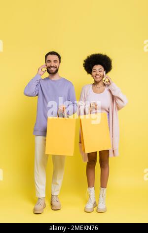 full length of happy interracial couple holding shopping bags and talking on smartphones on yellow background Foto Stock