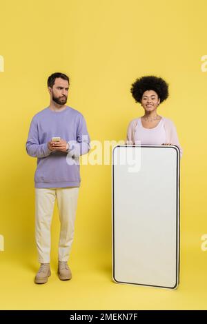 uomo barbuto con il telefono cellulare guardando afroamericano donna sorridente vicino cartoncino telefono mock-up su sfondo giallo Foto Stock