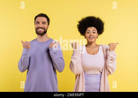 allegro coppia interracial che indica con i pollici e guardando la macchina fotografica isolata sul giallo Foto Stock
