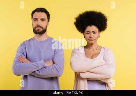 coppia interrazziale dispiaciuta in piedi con braccia incrociate mentre si guarda la macchina fotografica isolata sul giallo Foto Stock