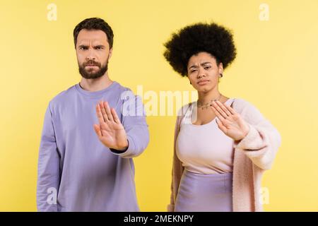 coppia multietnica dispiaciuta e accigliata che guarda la telecamera e mostra un gesto di rifiuto isolato sul giallo Foto Stock