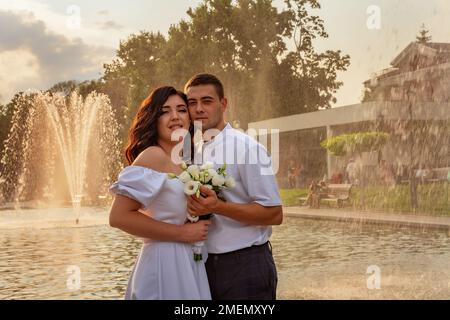 Coppia innamorata sullo sfondo di scintillanti spruzzi della fontana. Giovane coppia in abiti festosi con un bouquet di fiori festeggia Foto Stock