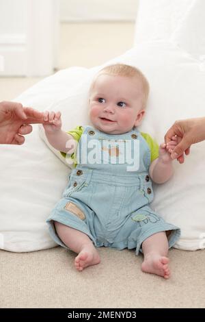 Due persone che tengono le mani del bambino (17 settimane) come si siede sostenuto da un cuscino grande Foto Stock