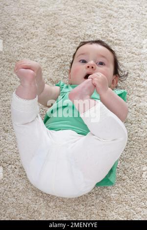 Bambina (6 mesi) sdraiata sul retro del pavimento con le gambe alzate tenendo i piedi Foto Stock