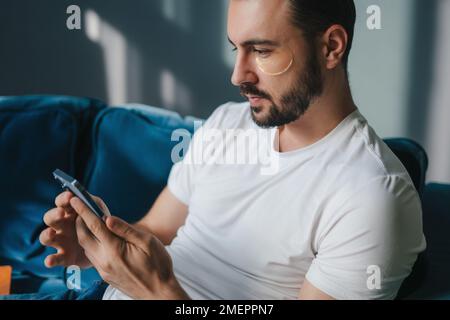 Uomo caucasico che usa lo smartphone mentre riposa su un divano nel soggiorno. Utilizzando i social network. Foto Stock