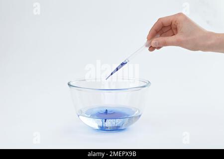 Utilizzando la pipetta, aggiungere gocce di vernice di seta blu all'acqua nel recipiente di vetro, primo piano Foto Stock