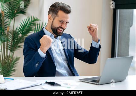 Felice uomo d'affari caucasico di successo in un vestito, ceo società, manager, siede alla scrivania in ufficio, gesturing con pugni, celebrare il successo, vincere, grande profitto, buon affare. Concetto di vittoria Foto Stock