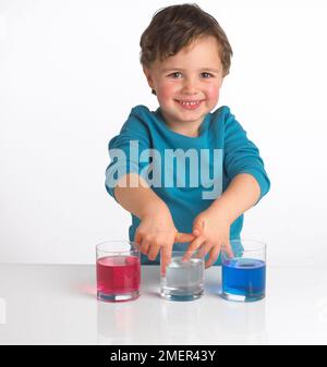 Ragazzo che indossa la parte superiore blu davanti a tre bicchieri contenenti liquido rosso, liquido trasparente e liquido blu, 4 anni Foto Stock