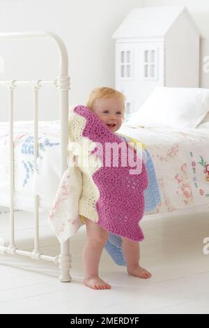 Bambino in piedi accanto al letto avvolto in coperta a maglia, 12 mesi Foto Stock