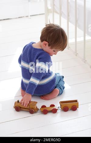 Ragazzo che indossa il ponticello uncinato che si inginocchia sul pavimento che spinge il giocattolo di legno, 2,5 anni Foto Stock