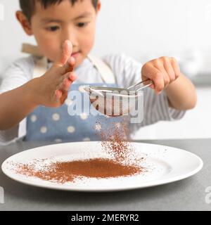 Ragazzo setacciando la polvere di cioccolato su un piatto, 4 anni Foto Stock
