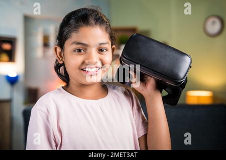 Immagine di una ragazza sorridente felice con VR o visore per realtà virtuale a portata di mano guardando la telecamera a casa - concetto di tecnologia, metaverse e. Foto Stock