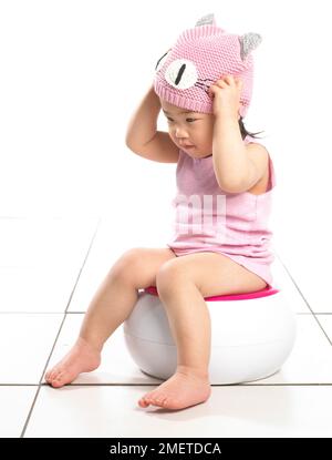 Ragazza che indossa giubbotto rosa seduta su vasino bianco, mettendo su cappello di lana rosa, 20 mesi Foto Stock