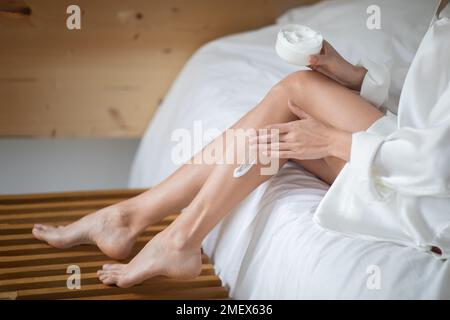 Giovane donna europea in accappatoio con gambe perfette siede su letto bianco con vaso di crema, applicare alla pelle Foto Stock