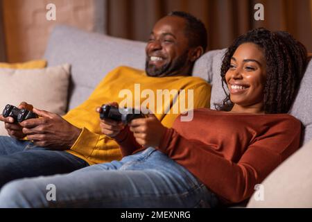Coniugi africani rilassati giocando a videogiochi a casa, vista laterale Foto Stock