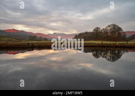 Splendida luce mattutina sulle montagne della Cumbria che si riflettono in un fiume calmo in una tranquilla mattinata sul fiume Brathay nel Lake District, Regno Unito. Foto Stock