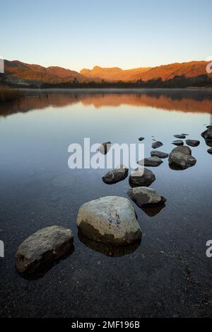 Luce mattutina sulle montagne di Elterwater nel Lake District, Regno Unito. Piccole rocce possono essere viste nel lago con riflessi di vetro nell'acqua. Foto Stock