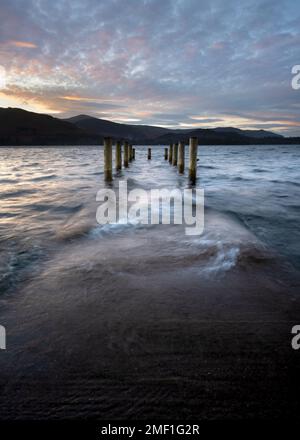 Molo in legno inondato con onde che si infrangono e che si affacciano su una spettacolare vista del tramonto sul lago. Derwentwater, Lake District, Regno Unito. Foto Stock