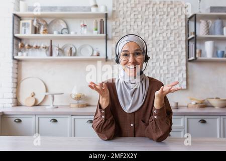 Donna hijab che lavora in remoto a casa, donna musulmana con cuffie per videochiamata sorridente e guardando la fotocamera, parlando con il cliente online in cucina, webcam vista pov. Foto Stock