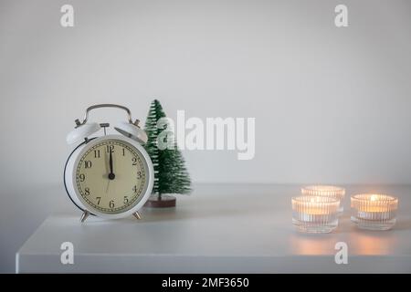 Una sveglia bianca, un puntatore a dodici, un piccolo albero di Natale artificiale verde e candele accese in candelabri su una cassettiera bianca. Copia Foto Stock