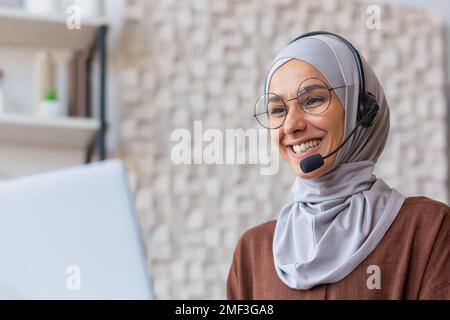 Donna allegra in hijab che lavora da casa in remoto, donna musulmana che parla in videochiamata con cuffia e notebook, donna d'affari che lavora da casa in remoto. Foto Stock
