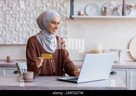 Donna allegra e di successo in hijab a casa in cucina con laptop e carta di credito banca effettua l'acquisto online in negozio online, donna musulmana sceglie prodotti e regali in remoto. Foto Stock