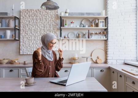 Freelancer in hijab lavorando sul laptop in remoto da casa, donna ha ricevuto buone notizie e buone notizie on-line notifica, felice e celebrando i buoni risultati di realizzazione, donna d'affari in casa ufficio. Foto Stock