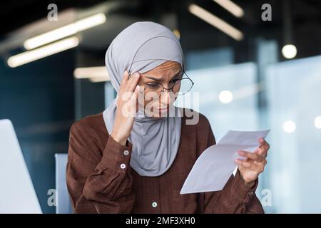 Primo piano. Una giovane araba preoccupata in un hijab siede in ufficio e tiene una lettera, un documento nelle sue mani. Tiene la testa, ha ricevuto cattive notizie, divorzio, debiti, fallimento. Foto Stock