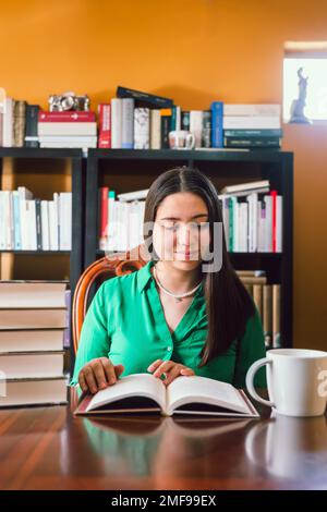 Giovane donna che legge il libro nella sua biblioteca personale con libreria dietro di lei, con una tazza di caffè. Foto Stock
