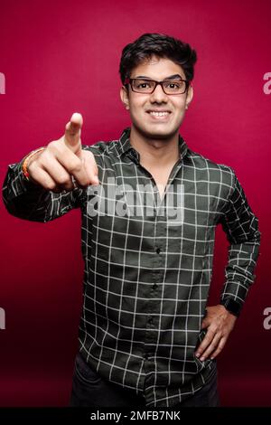 Ritratto di giovane indiano sorridente in camicia a scacchi e occhiali che puntano la macchina fotografica su sfondo rosso Foto Stock