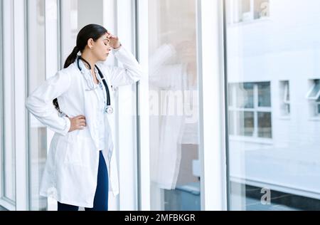 I medici hanno uno dei lavori più stressanti del mondo. una giovane donna che guarda stressato fuori mentre in piedi in una finestra in un ospedale. Foto Stock