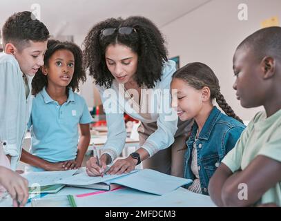 Insegnante femminile in classe con i bambini, aiutando la studentessa con il lavoro scolastico e scrivendo nel libro. Diversità nell'istruzione, educatore che legge i capretti Foto Stock