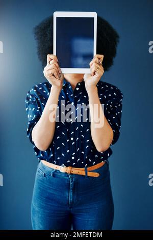 Consente di rendere visibile la tua app. Ripresa in studio di una giovane donna che ricopre il viso con un tablet digitale vuoto su sfondo blu. Foto Stock