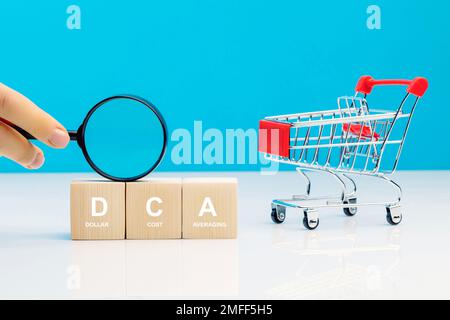 Lente d'ingrandimento in mano della donna e parola DCA su un cubo di legno in idea Dollaro costo Averaging strategia d'investimento vicino carrello vuoto. Salvataggio della stoc Foto Stock