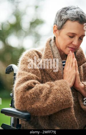 La sua fede che la mantiene andare. una donna anziana che prega mentre si siede in sedia a rotelle all'aperto. Foto Stock