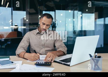 Concentrato giovane afroamericano maschio ragioniere, auditor, banchiere che lavorano in ufficio alla scrivania con computer portatile e documenti. Firma accordi, scrive documenti. Foto Stock