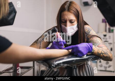 Tatuaggio artista tatuaggio nel suo studio con guanti e maschera. Braccio del tatuaggio. Foto Stock