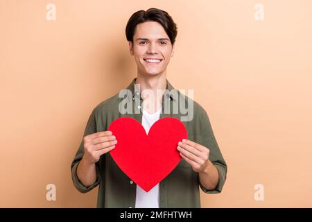 Ritratto foto di giovane ottimista bel ragazzo brunetto capelli tenere grande amore rosso simbolo come la sua ragazza dono isolato su sfondo di colore beige Foto Stock