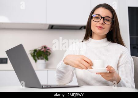 Ritratto di una giovane bella donna che tiene una tazza di caffè e seduto in cucina vicino al computer portatile. Il freelance ha una pausa Foto Stock