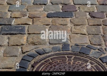 Vecchio portello della fogna del metallo nel mezzo della strada, disposto da una pietra di pavimentazione liscia. Foto Stock