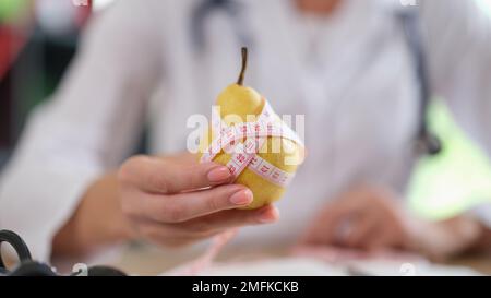 Nutrizionista che mostra pera gialla matura con metro a nastro mentre si siede in ufficio medico. Foto Stock