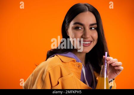 Bella giovane donna che tiene bere paglia in bottiglia con succo isolato su arancione, immagine stock Foto Stock