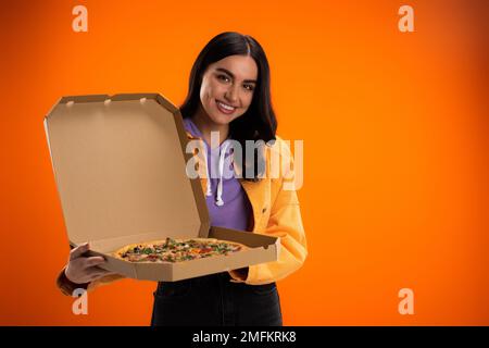 donna gioiosa che guarda la macchina fotografica mentre tiene scatola di cartone con gustosa pizza isolata su arancione, immagine stock Foto Stock