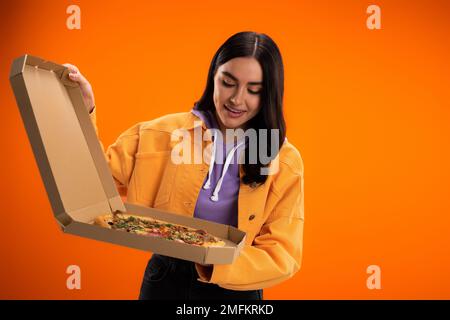 donna compiaciuta in giacca alla moda guardando gustosa pizza in scatola di cartone isolato su arancione, immagine stock Foto Stock