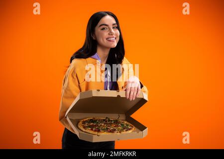 donna sorridente brunette che mostra gustosa pizza in scatola di cartone mentre guarda la fotocamera isolato su arancione, immagine stock Foto Stock