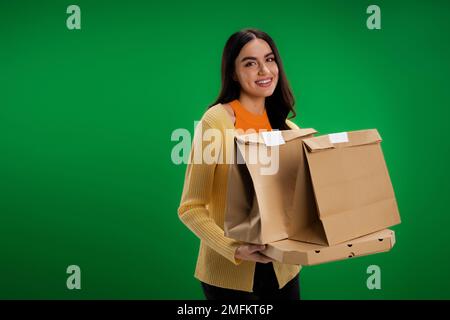 felice donna brunette che tiene sacchetti di carta e scatola di pizza mentre guarda la macchina fotografica isolata sul verde, immagine di scorta Foto Stock