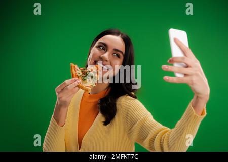 allegra donna mordente pezzo di pizza e prendendo selfie su smartphone isolato su verde, immagine stock Foto Stock